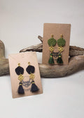 Close-up of black and green Mizuhiki plum knot earrings with matal flower charm and small tassel, handmade by Itookasi. A blend of Japanese craft and Finnish nature symbolizing strong bonds.